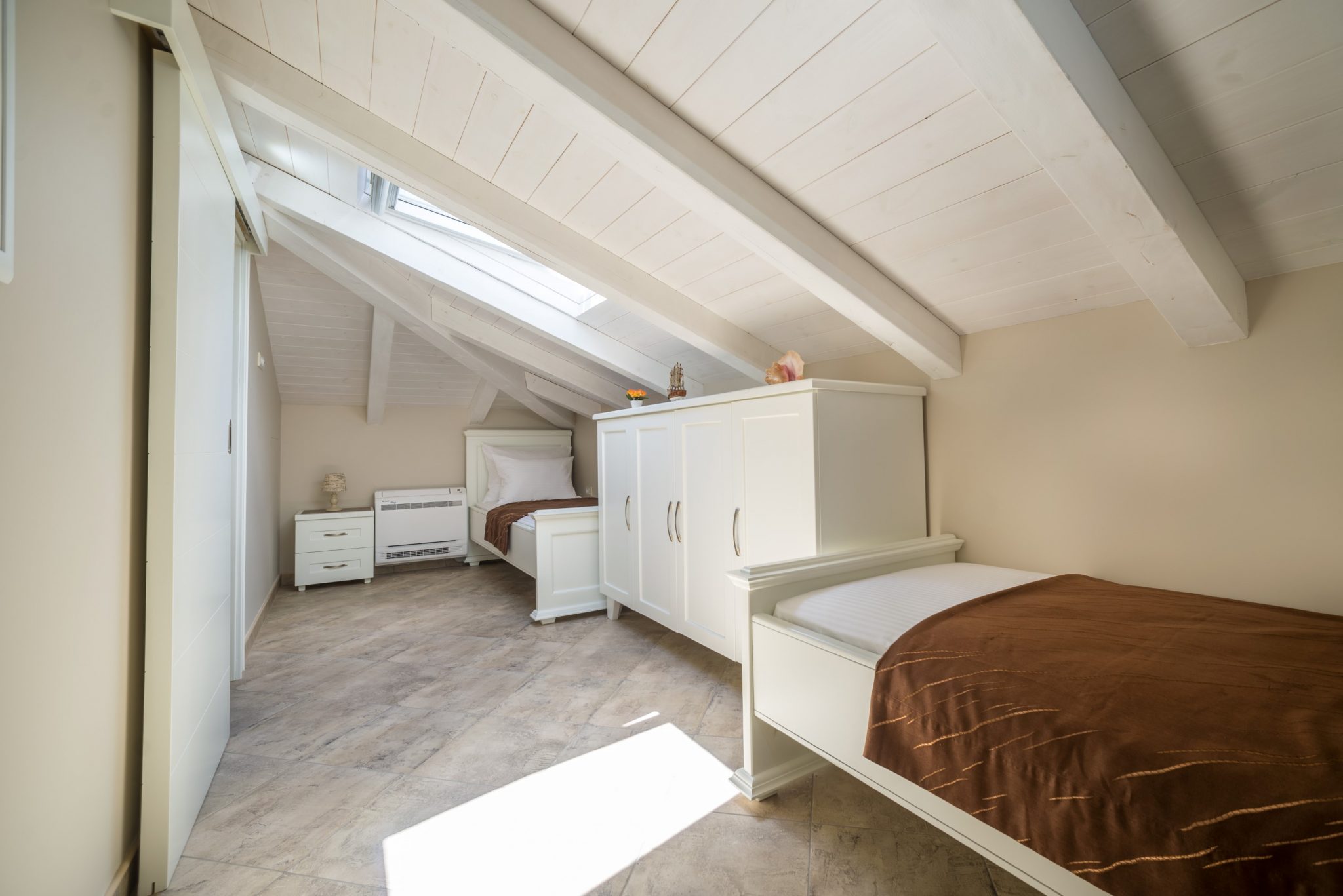 Attic bedroom with white furniture and a sloped ceiling in a villa in Stoliv, Montenegro. Two beds and a wardrobe.