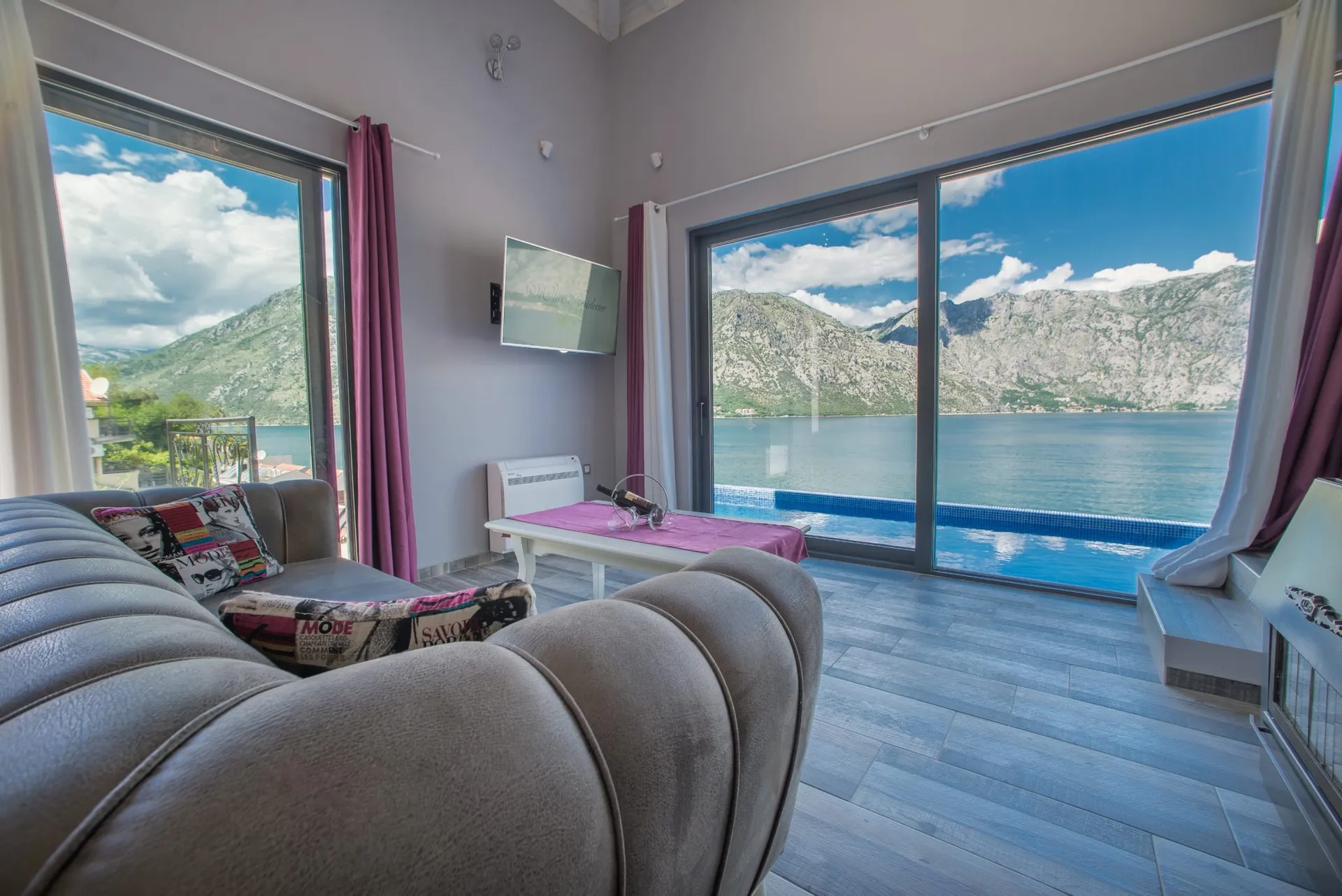 Living room with a grey sofa, wall TV, and large windows overlooking the sea and mountains.
