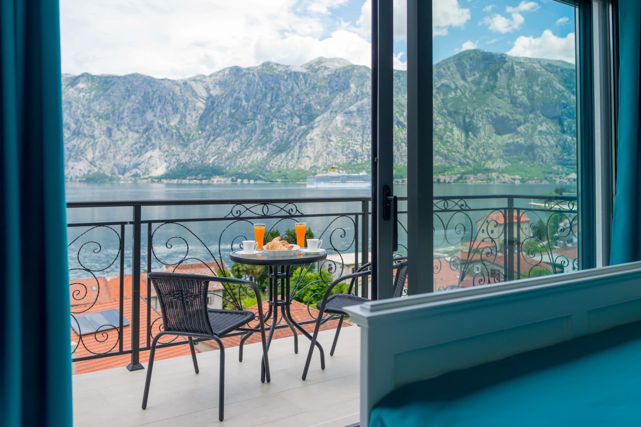 Villa balcony in Stoliv, Montenegro: view of the Bay of Kotor, mountains, and a cruise ship, with a breakfast table.