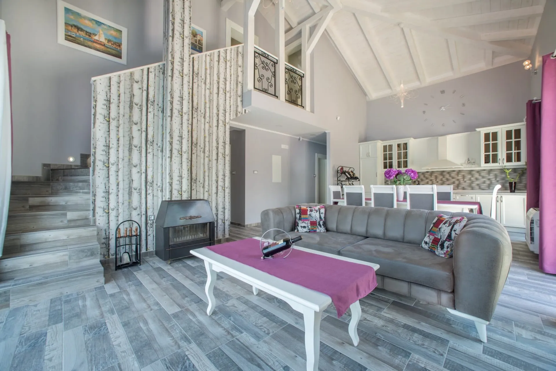 Living room with grey sofa, fireplace, and birch-patterned staircase wall, kitchen visible in background.