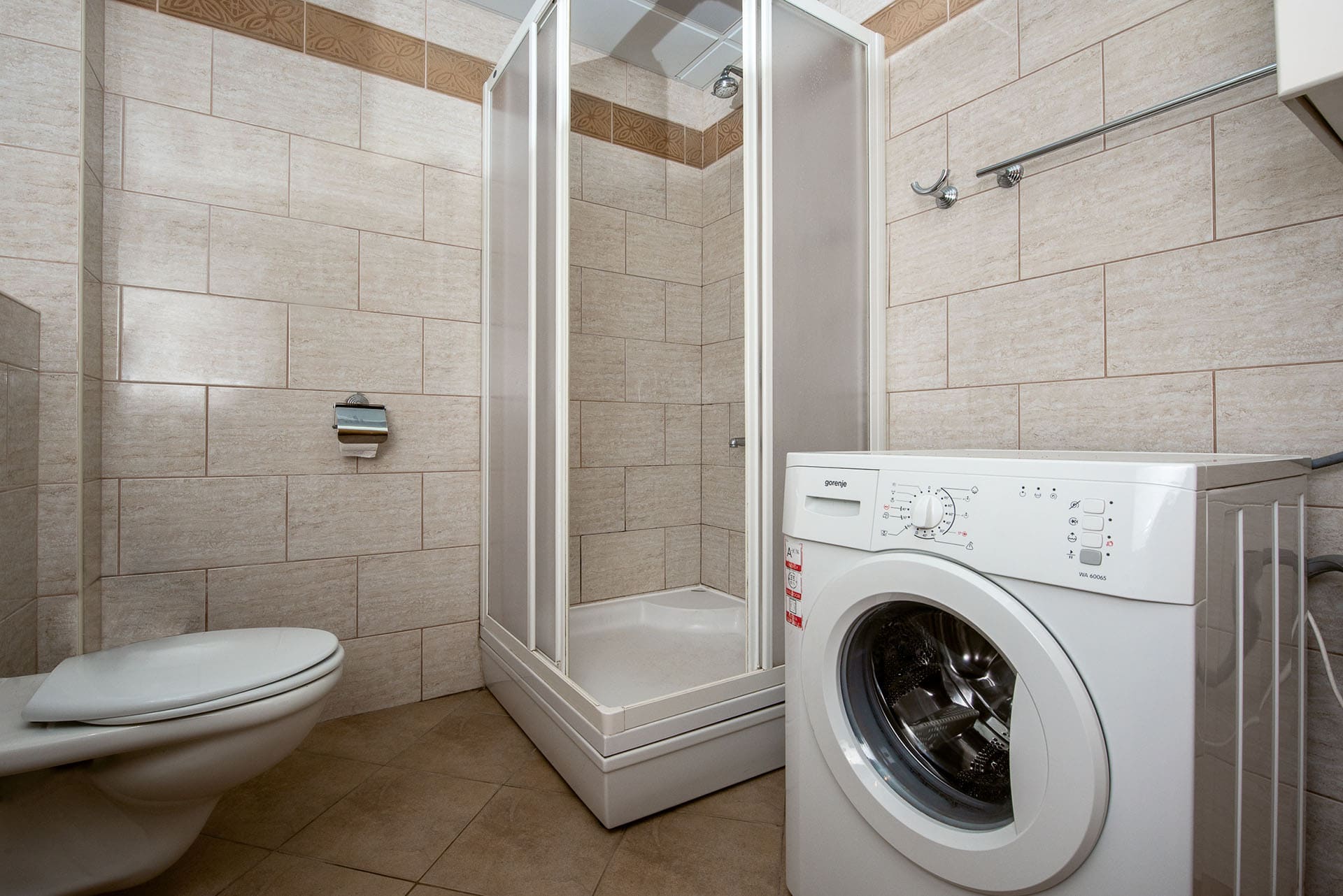 Bathroom in an apartment in Bečići, Montenegro: shower cabin, toilet, and Gorenje washing machine, beige tiles.