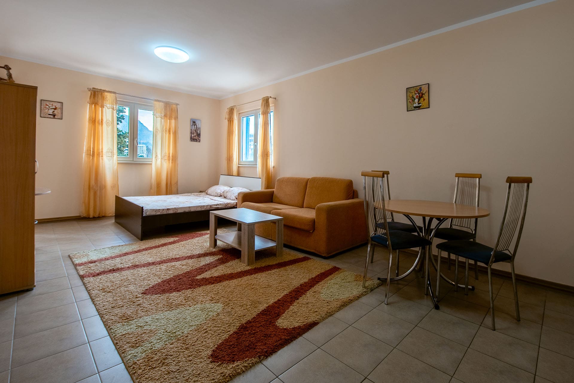 Living room in an apartment in Bečići, Montenegro: a double bed, an orange sofa, a dining table, and tiled flooring.