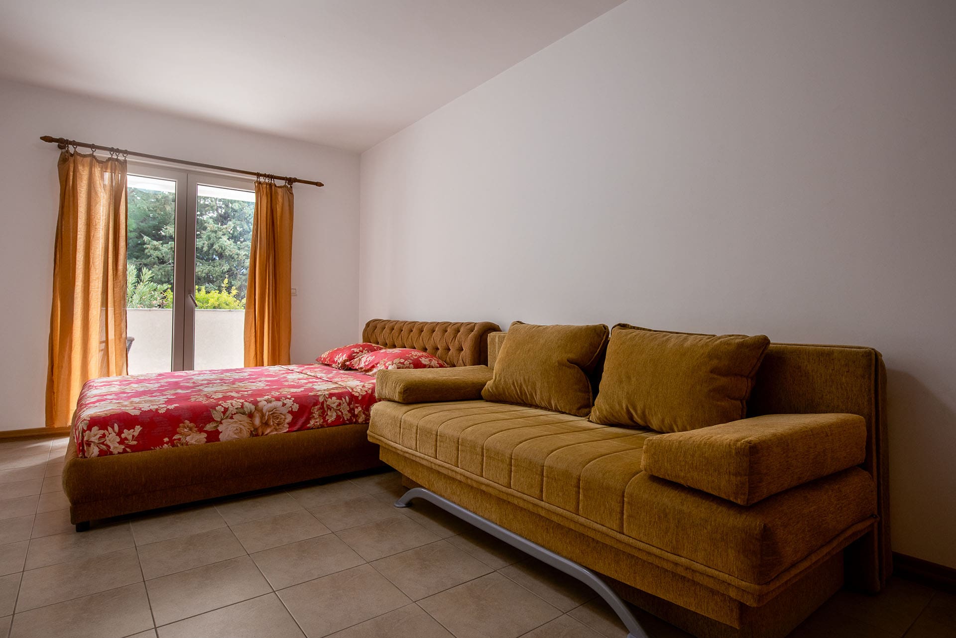 Bedroom in an apartment in Bečići, Montenegro: a double bed, a mustard-colored sofa, and access to a balcony overlooking greenery.