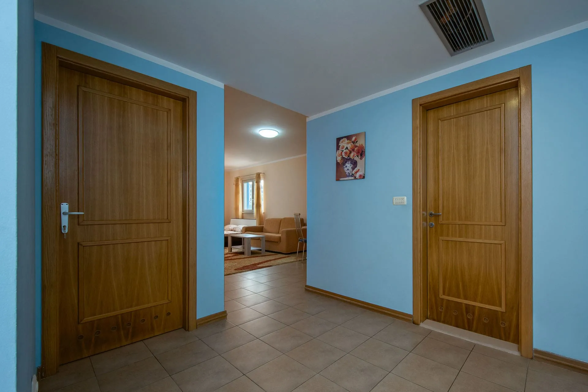 Bright hallway with two wooden doors and tiled floor. A living room with a sofa is visible through the central doorway.
