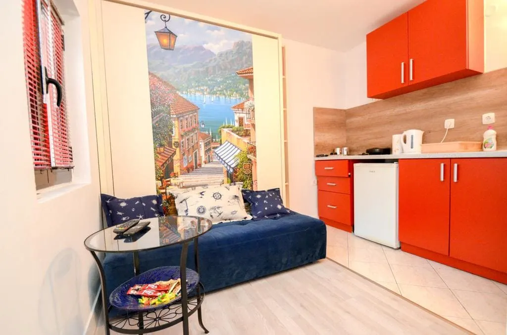Small room with a blue sofa, glass table, and orange kitchen area with cabinets.