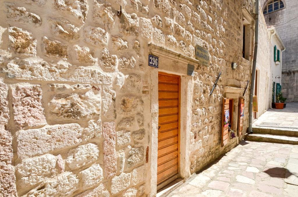 Facade of an old stone house in the historic center of Kotor, Montenegro, with a wooden door and cobblestone paving.
