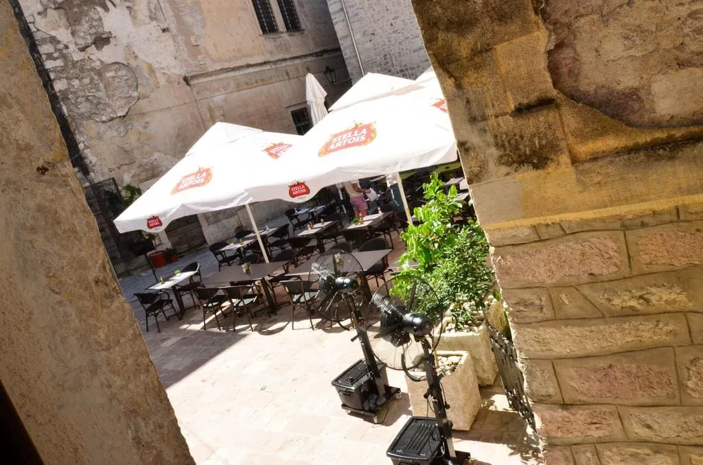 Outdoor cafe terrace with white umbrellas, tables, and fans set against an old stone wall.