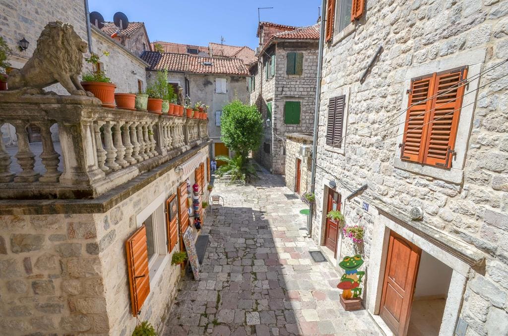 A narrow cobblestone street in the Old Town of Kotor, Montenegro, with stone houses, shutters, and a terrace with a lion statue.