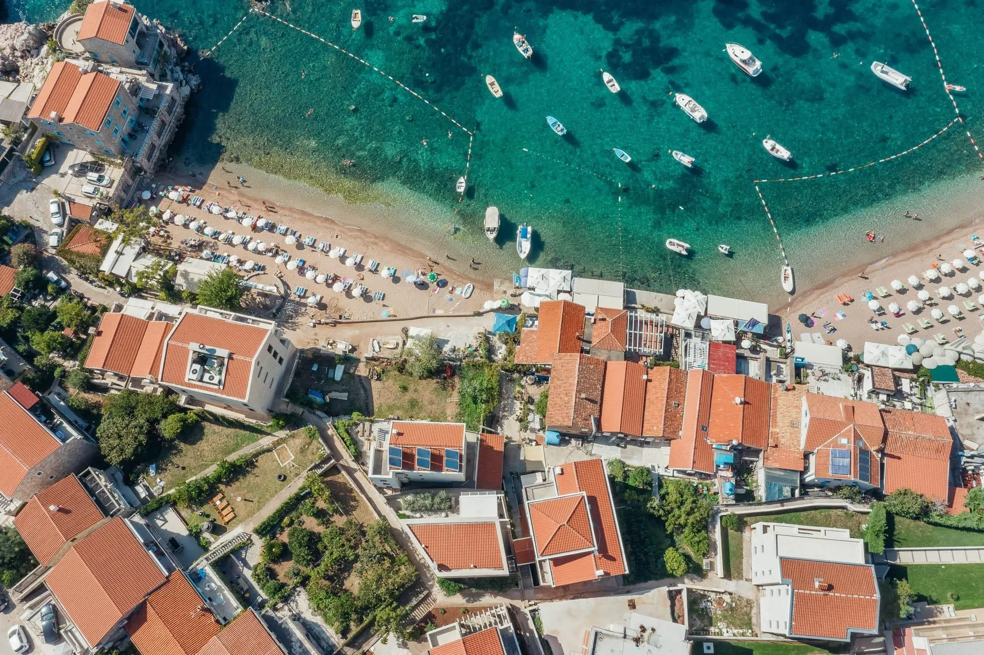 Aerial view of a coastal village with red-tiled roofs, a sandy beach lined with umbrellas, and boats in turquoise water.