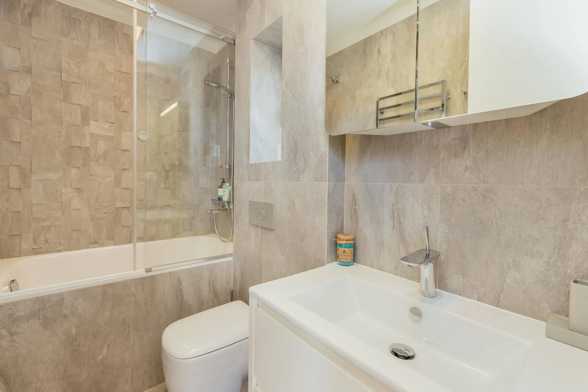 Bathroom featuring a bathtub with shower screen, white toilet, vanity sink, and mirrored cabinet against beige tiled walls.