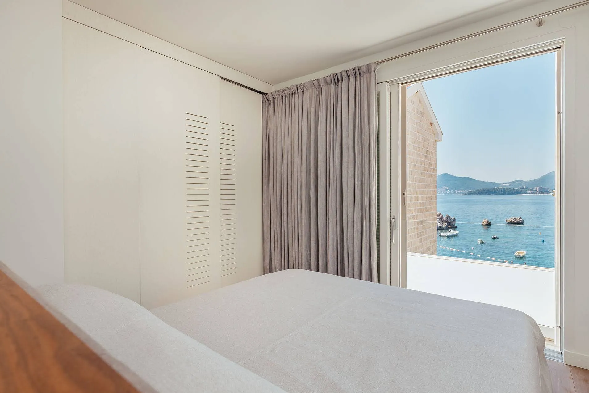 Bright bedroom with a white bed and built-in wardrobe. The open door reveals a scenic view of the sea, rocks, and boats.
