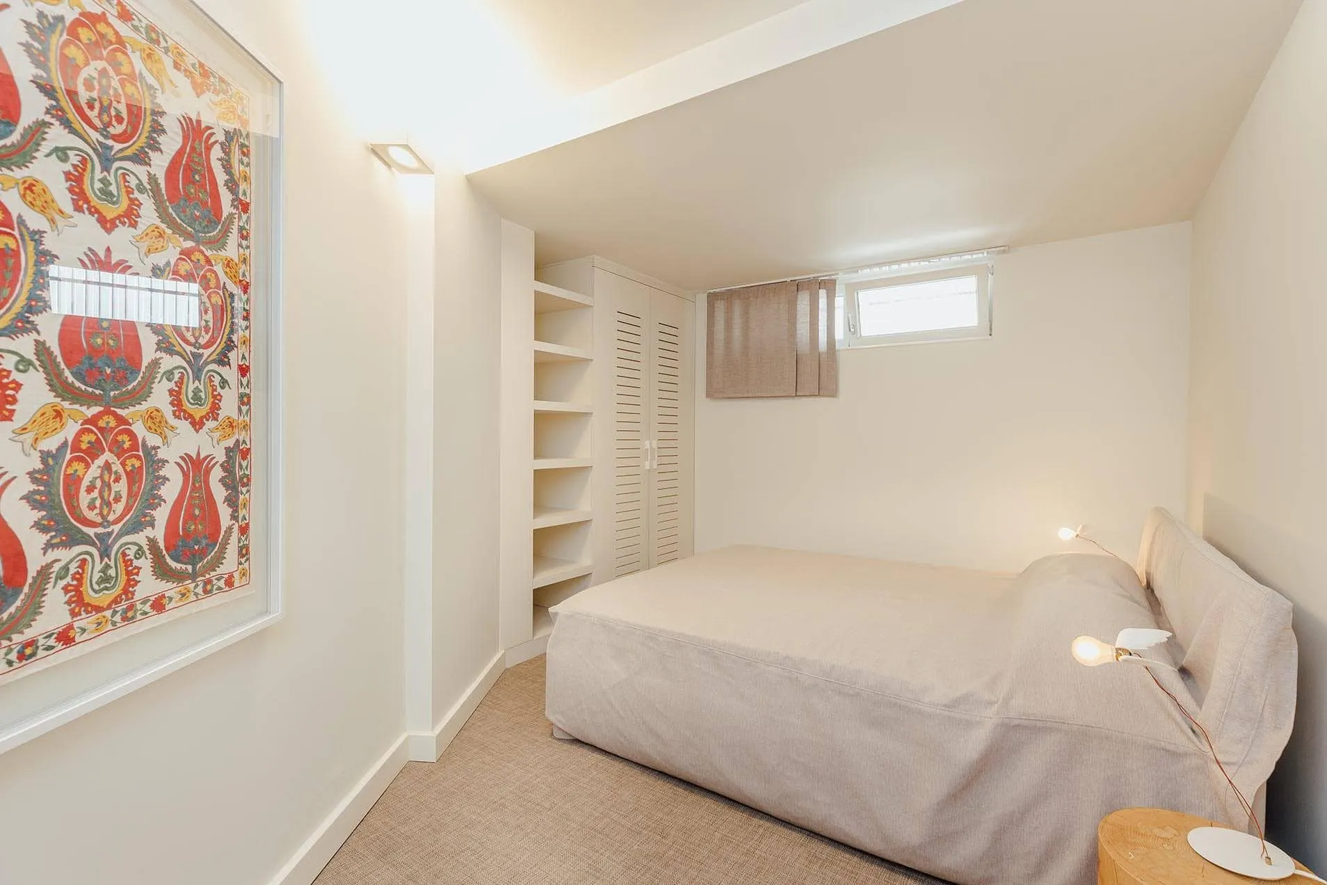 Bright bedroom featuring a single bed, built-in white wardrobe with shelves, and a colorful textile poster on the wall.