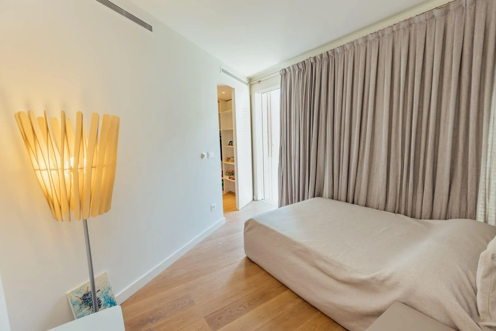 Bright bedroom with wooden floors, a beige mattress on the floor, and a tall slatted wood lamp against a white wall.