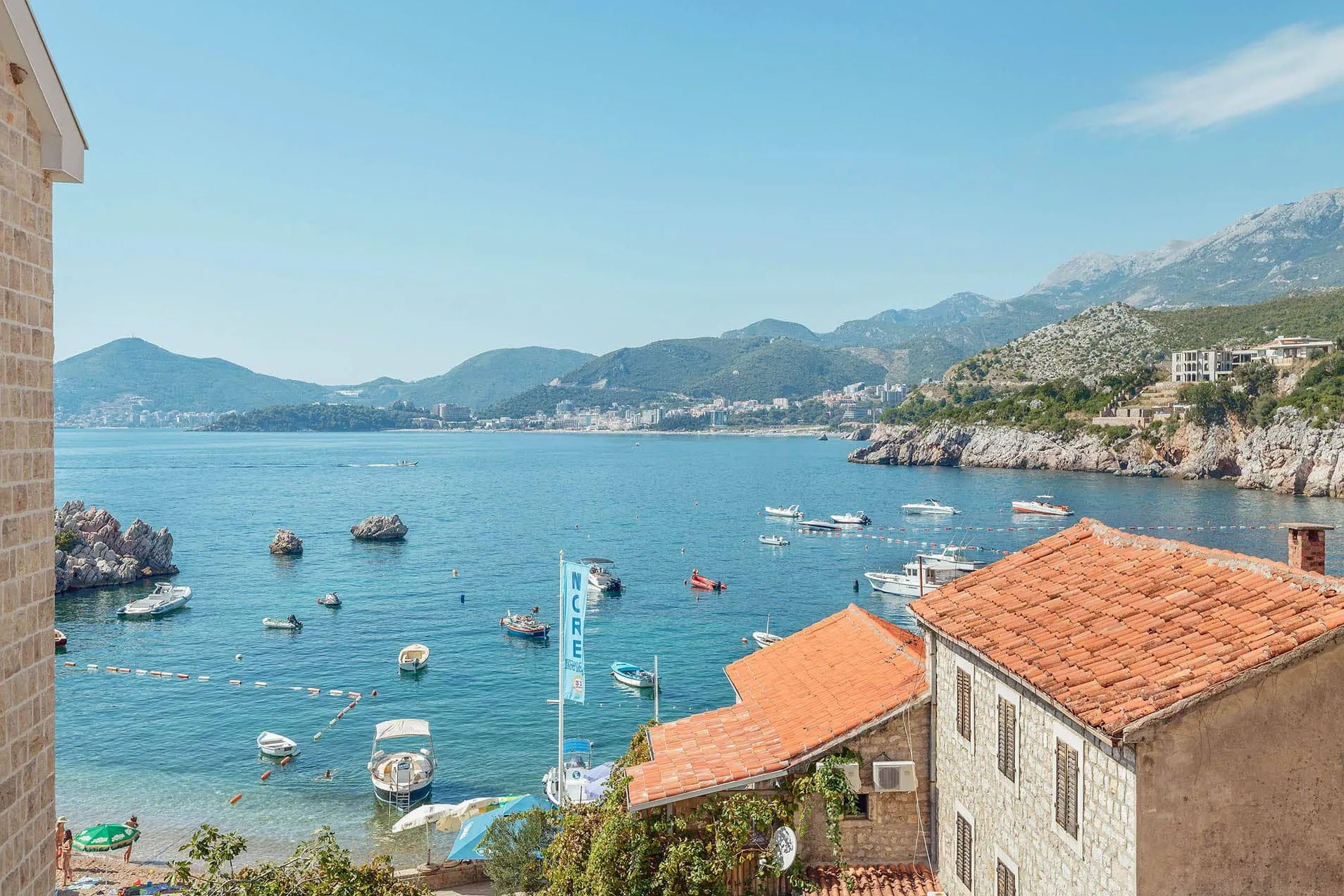 Panoramic view of a sea bay with boats, stone houses with red tiles, and mountains in the background under a blue sky.