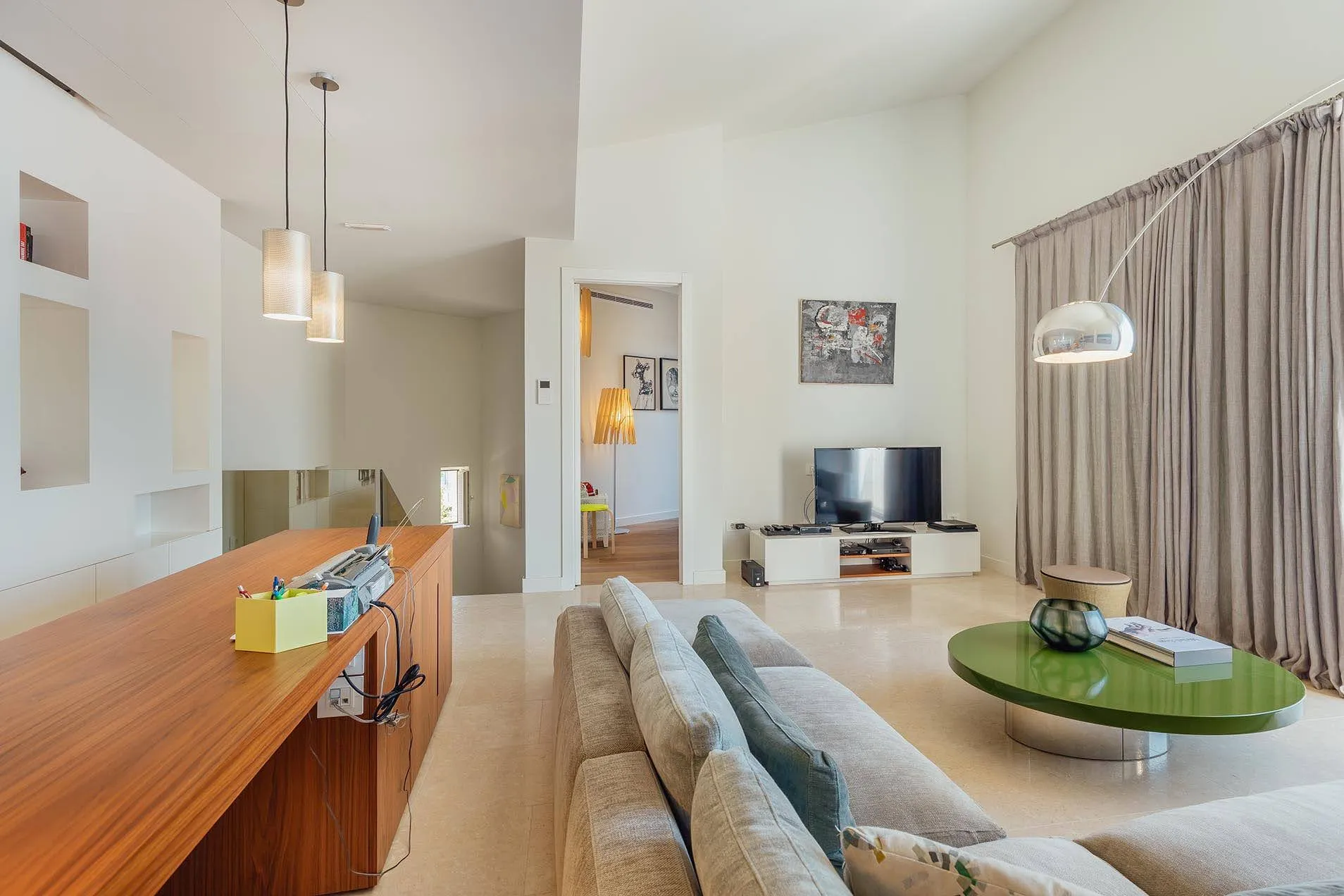 Bright room with a gray sofa, green table, TV, and wooden counter featuring pendant lights.