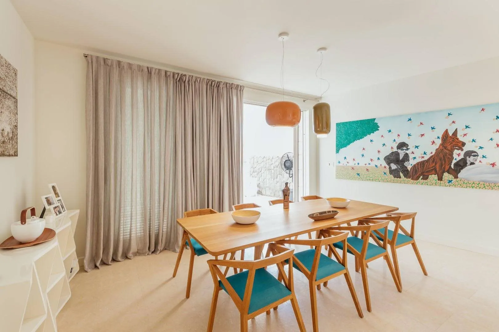 Bright room with a wooden dining table, six chairs with teal seats, pendant lights, and a large colorful wall mural.