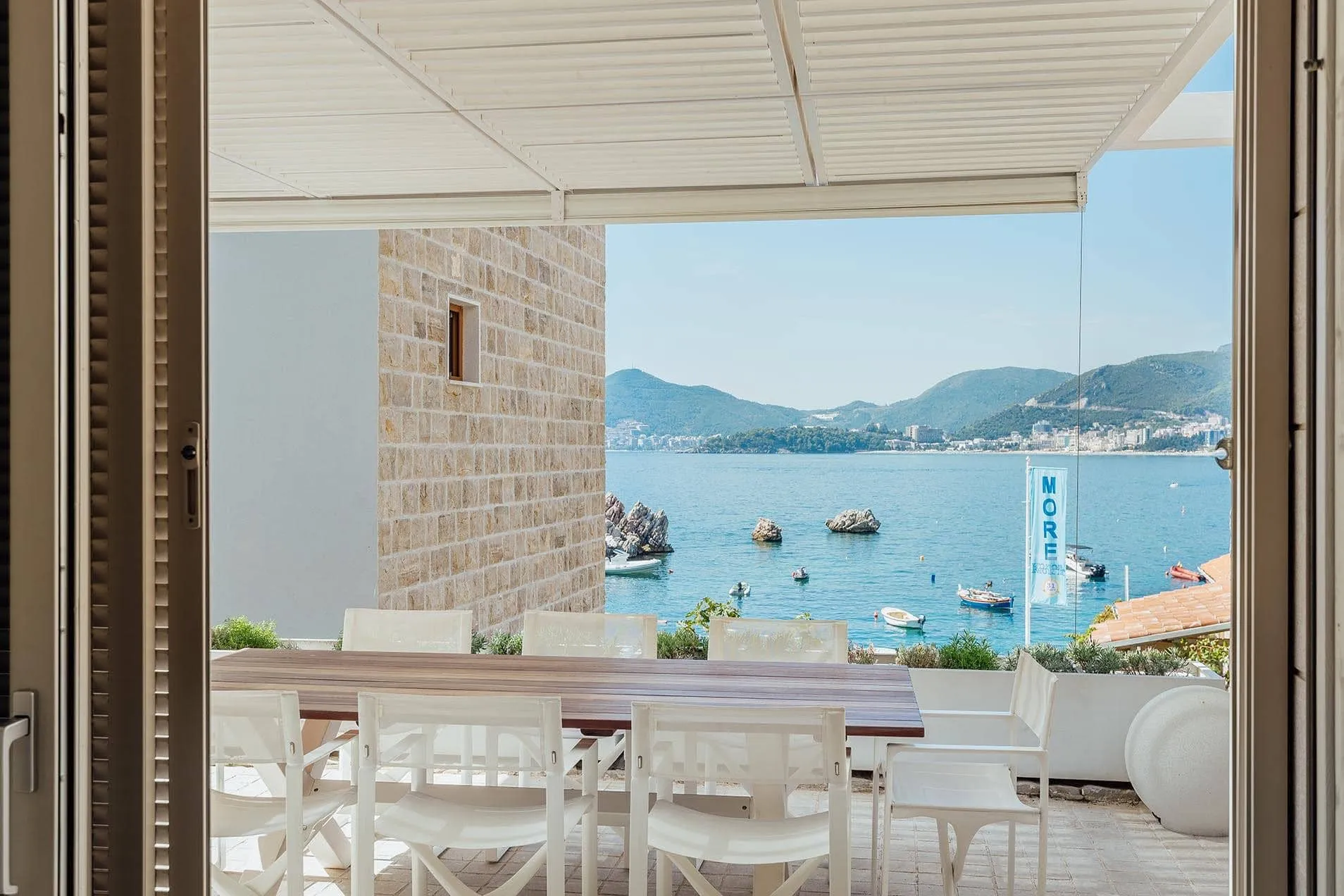 Balcony with white chairs and wooden table under a pergola, overlooking the sea, rocks, and boats.