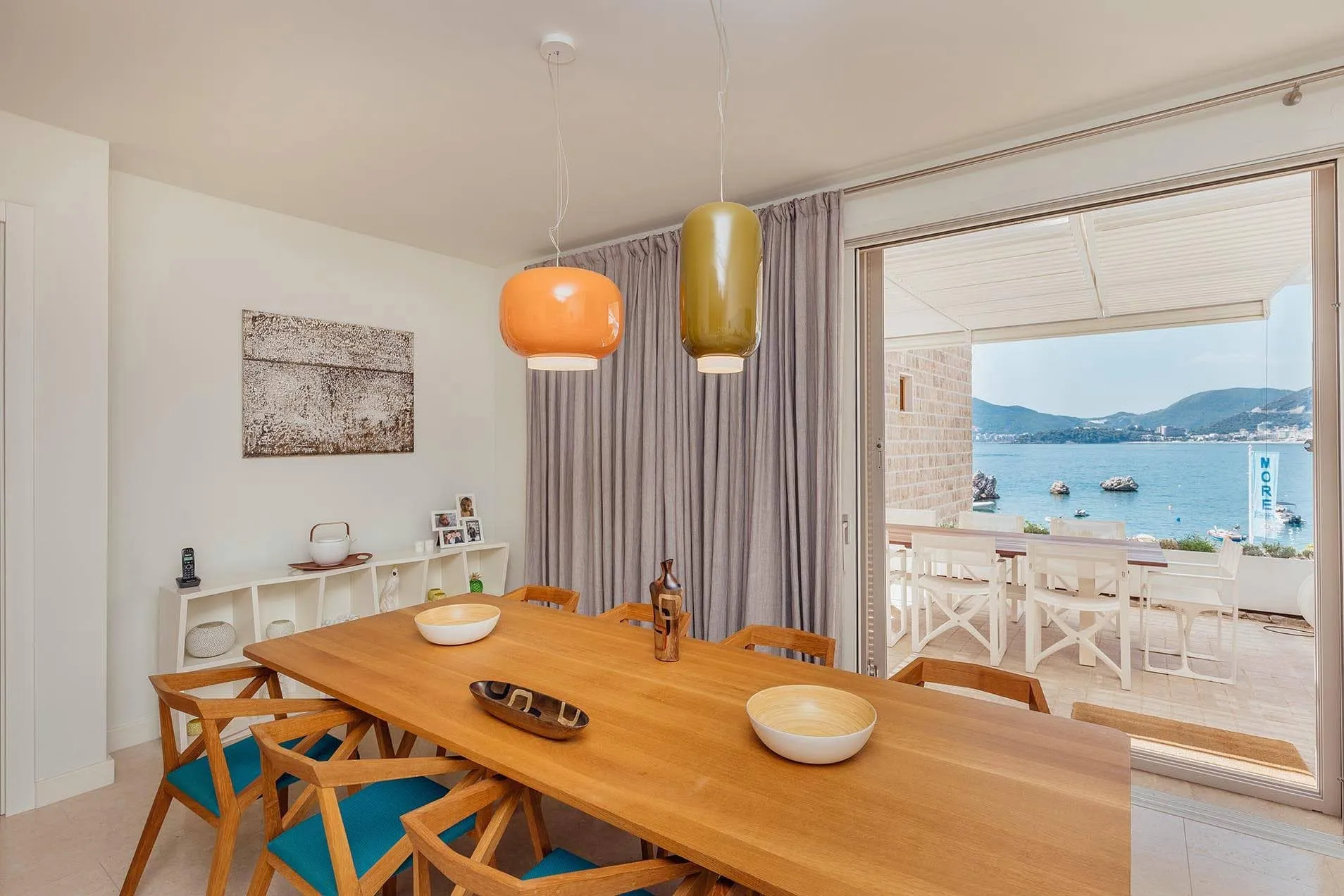 Dining area with a wooden table and colorful pendant lamps, leading to a balcony with a sea view.