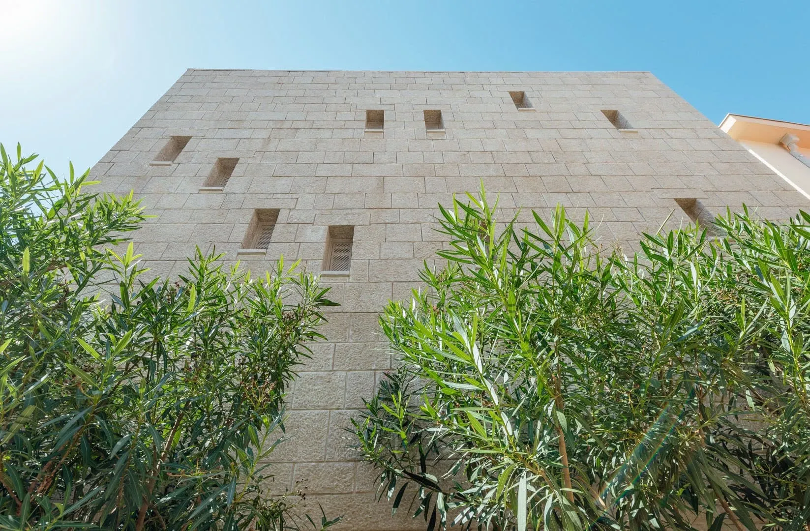 View of a stone building facade with small windows and green bushes in the foreground under a clear blue sky.
