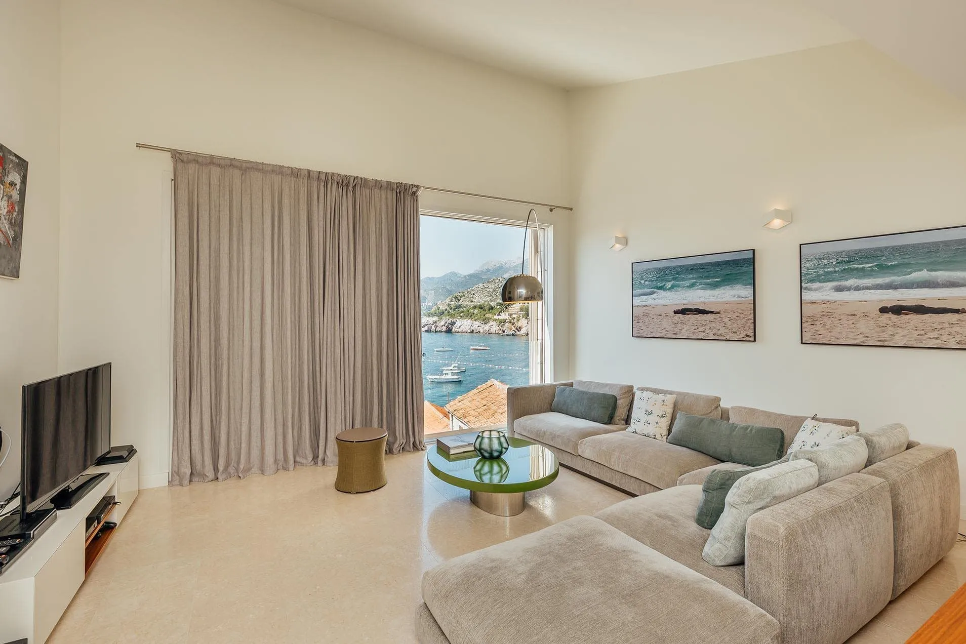 Bright living room with a beige sectional sofa, TV unit, and round glass table. Large window shows a sea view.