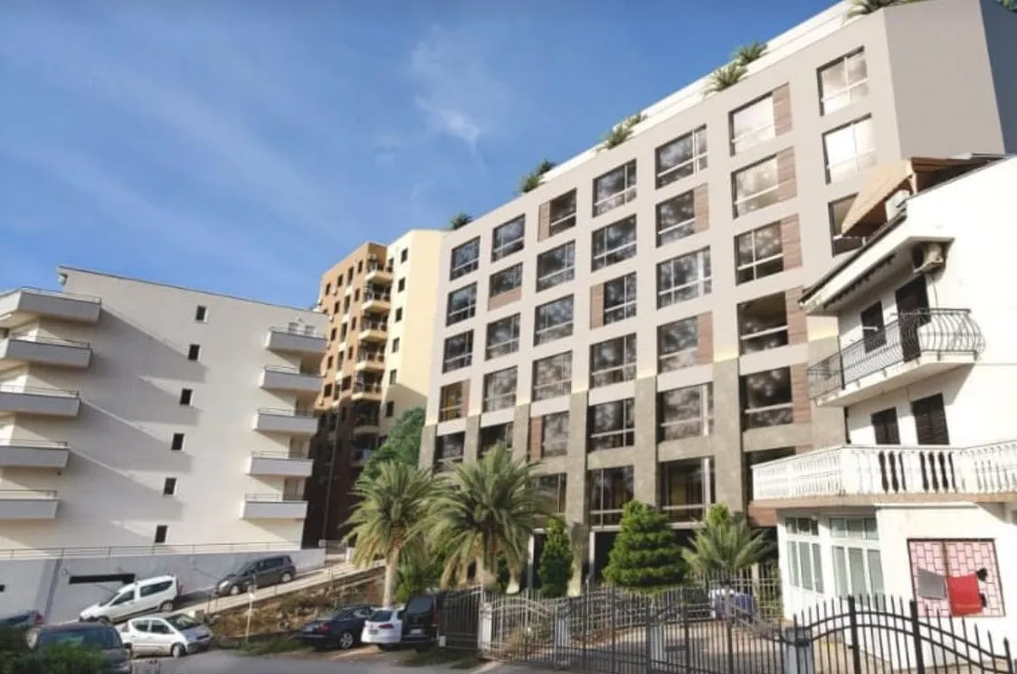 Exterior view of multi-story residential buildings with balconies, palm trees, and parked cars under a blue sky.