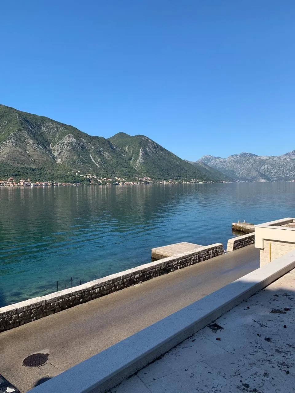 Seascape with a stone embankment, mountains, and a village across the water under a clear blue sky.