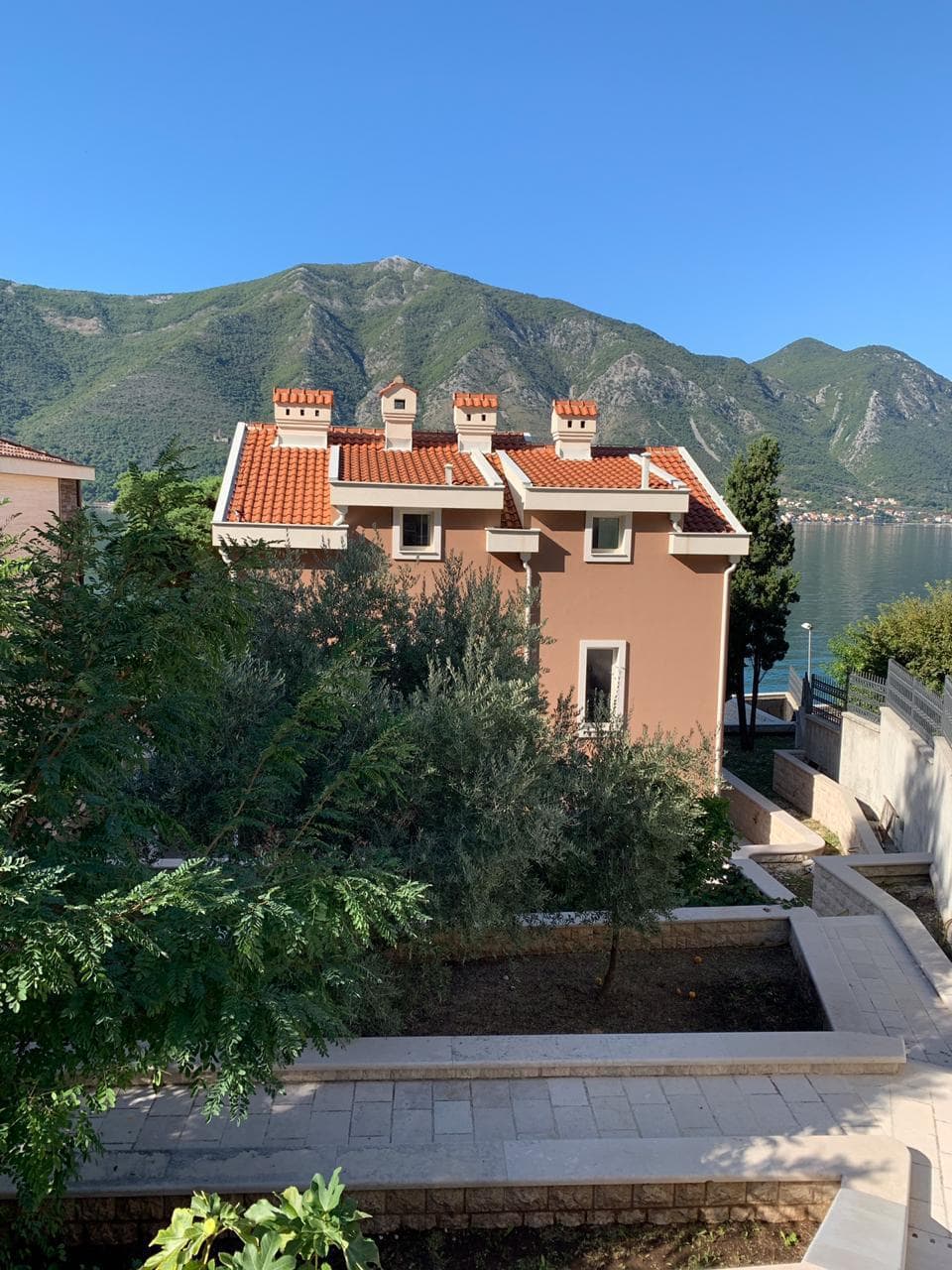 Villa with a tiled roof in Dobrota, Montenegro: view of the house, green garden, and sea bay with mountains in the background.