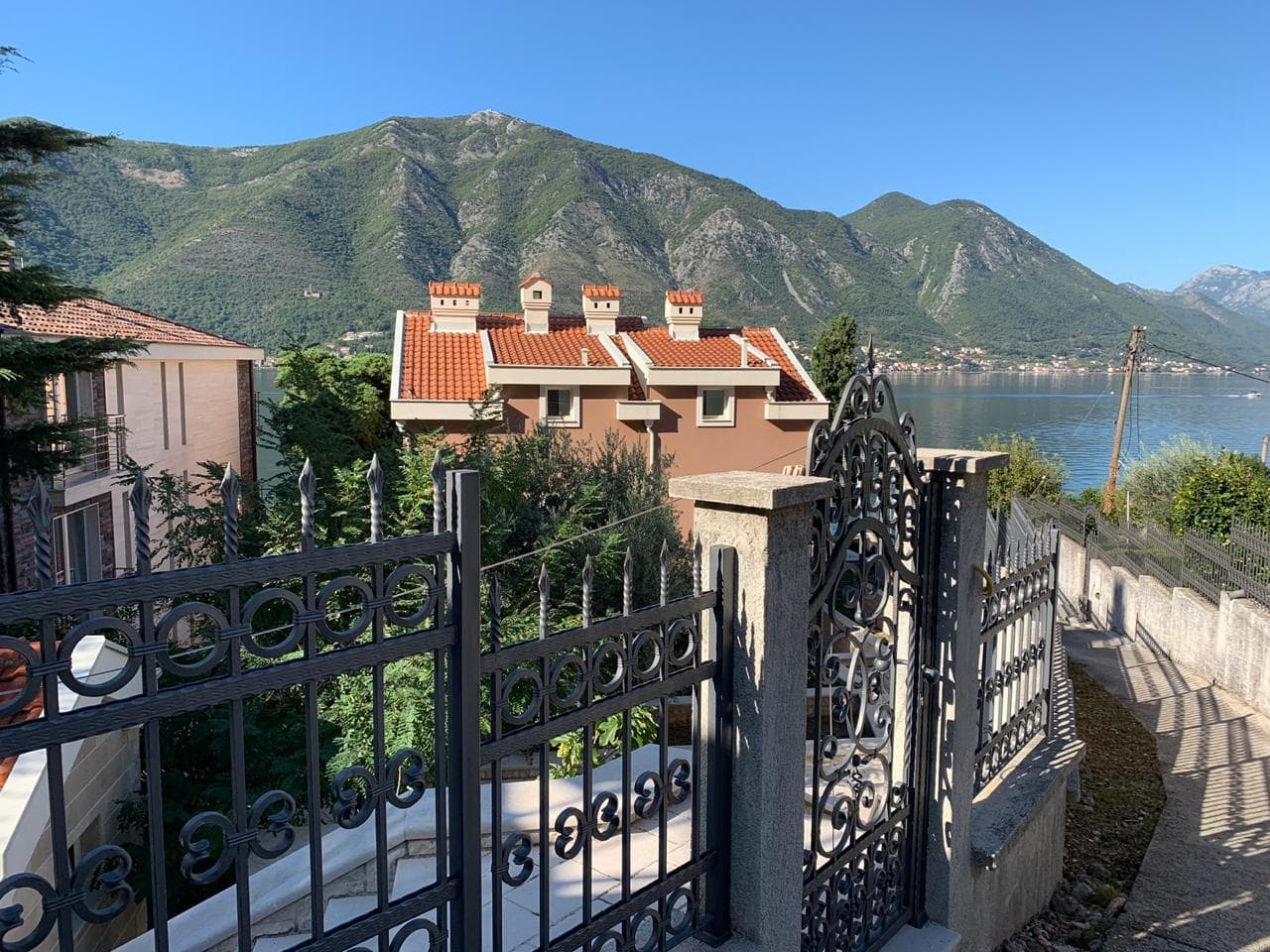 View of a villa in Dobrota, Montenegro: wrought-iron fence, tiled roof, and panoramic views of the Bay of Kotor and the mountains.
