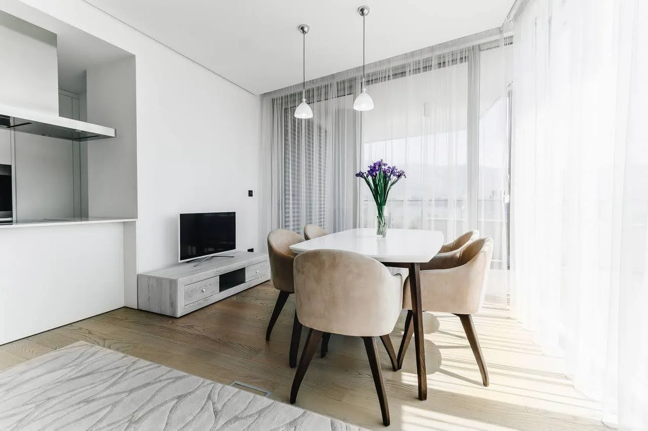 Bright room with a white dining table, beige chairs, TV on a low console, and an open kitchen area visible on the left.