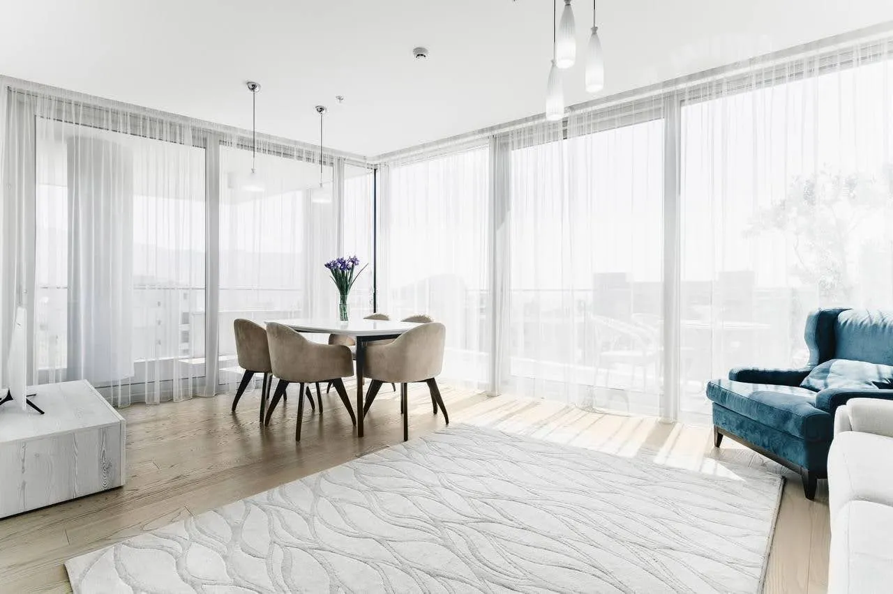 Bright room with a dining set, blue sofa, white rug, and floor-to-ceiling windows covered by sheer curtains.