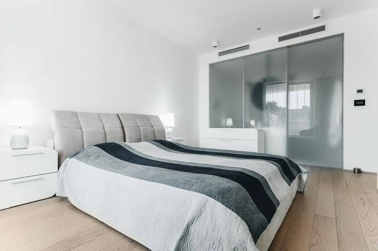 Modern bedroom featuring a double bed with striped bedding, white nightstands, and mirrored wardrobe doors.