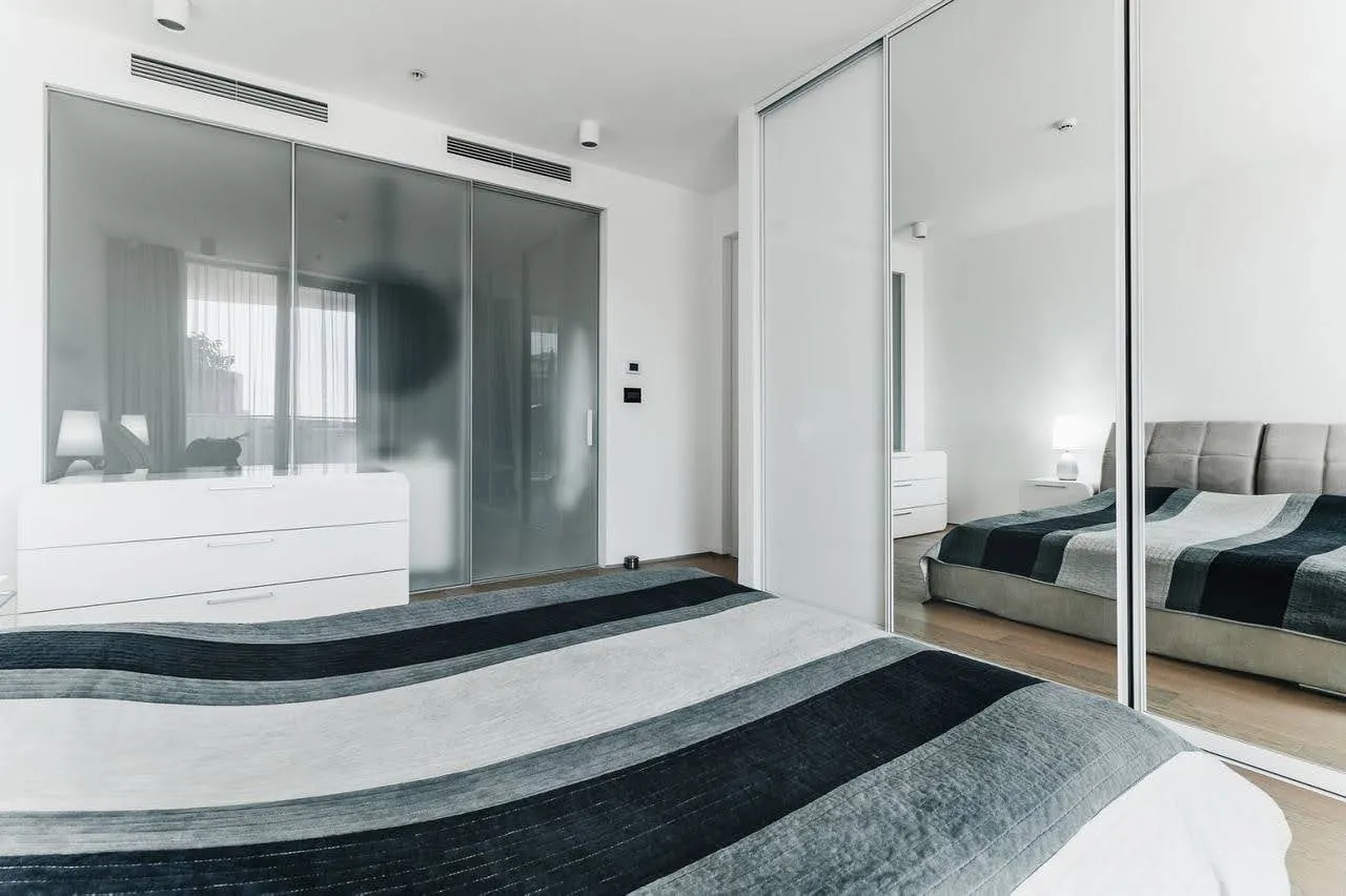 Modern bedroom featuring a bed with striped cover and white dresser next to glass panels.