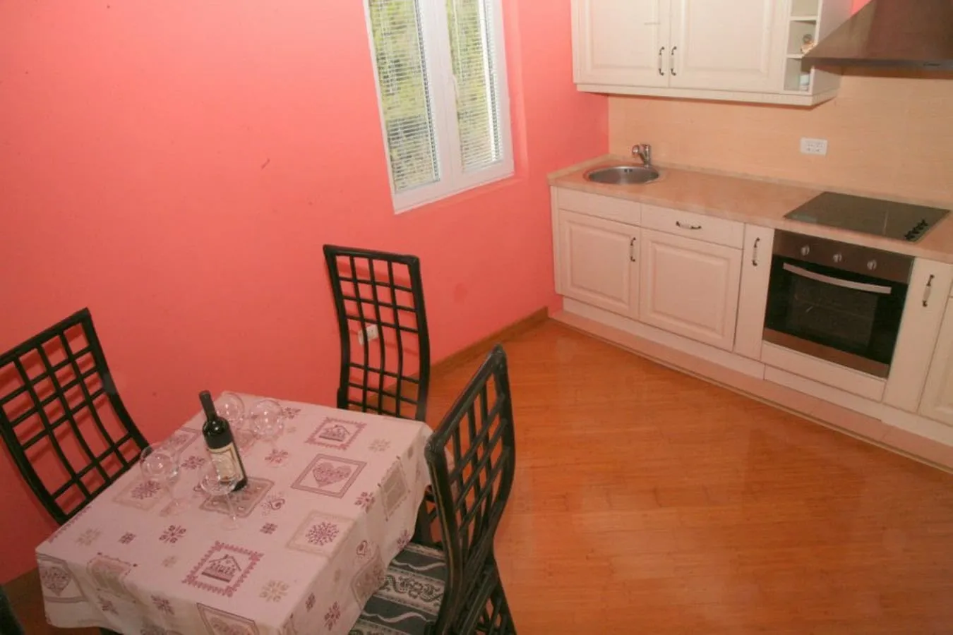 Kitchen with white cabinets, sink, stove, and oven. Pink walls, wooden floor, dining table with black chairs and wine bottle.