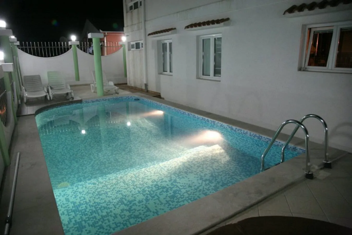 Outdoor swimming pool with blue mosaic tiles and underwater lights at night, surrounded by lounge chairs.