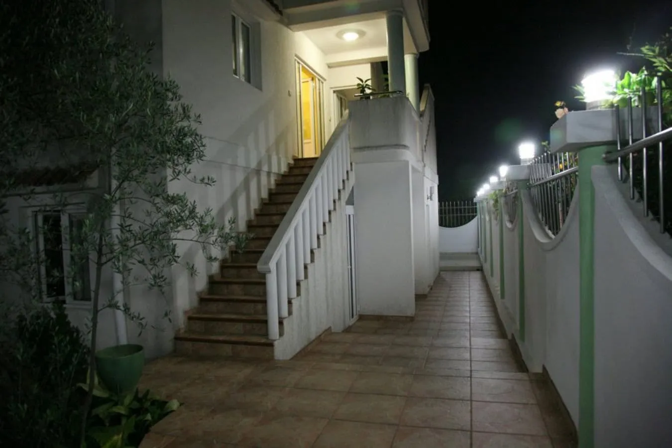 Night view of an outdoor staircase and tiled walkway alongside a white house with lights and plants.