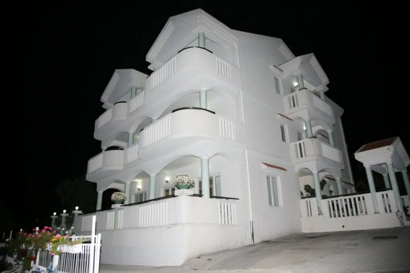 Night view of a white multi-story building with balconies, columns, and flower boxes against a black sky.