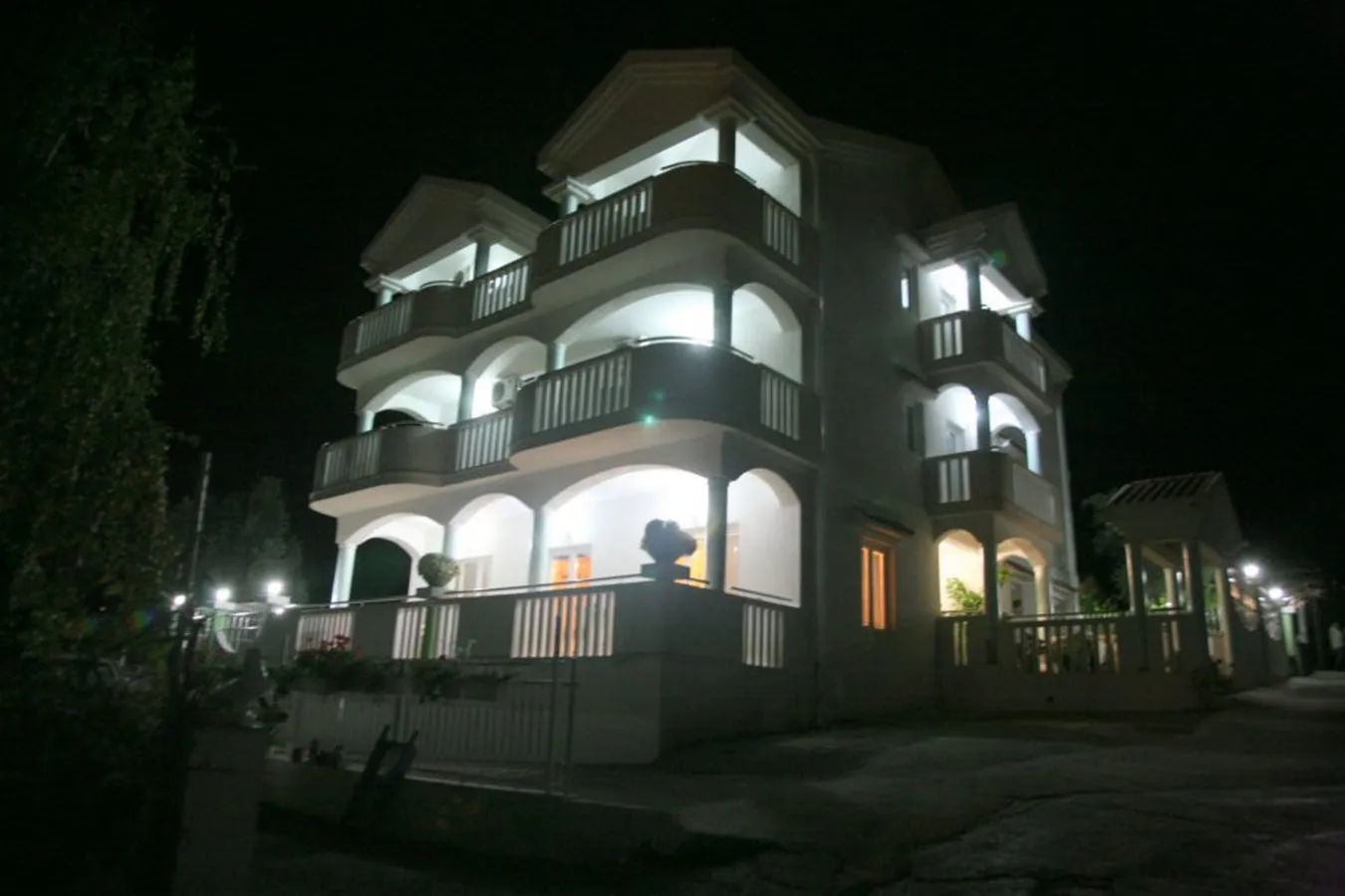 Night exterior view of a multi-story white house with balconies and arches, illuminated by outdoor lights.