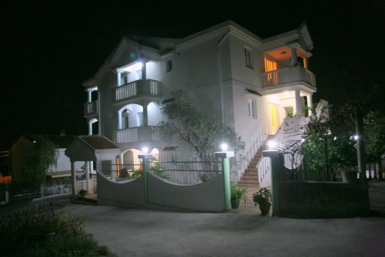 Night view of a two-story house with balconies, illuminated stairs, and a perimeter fence.