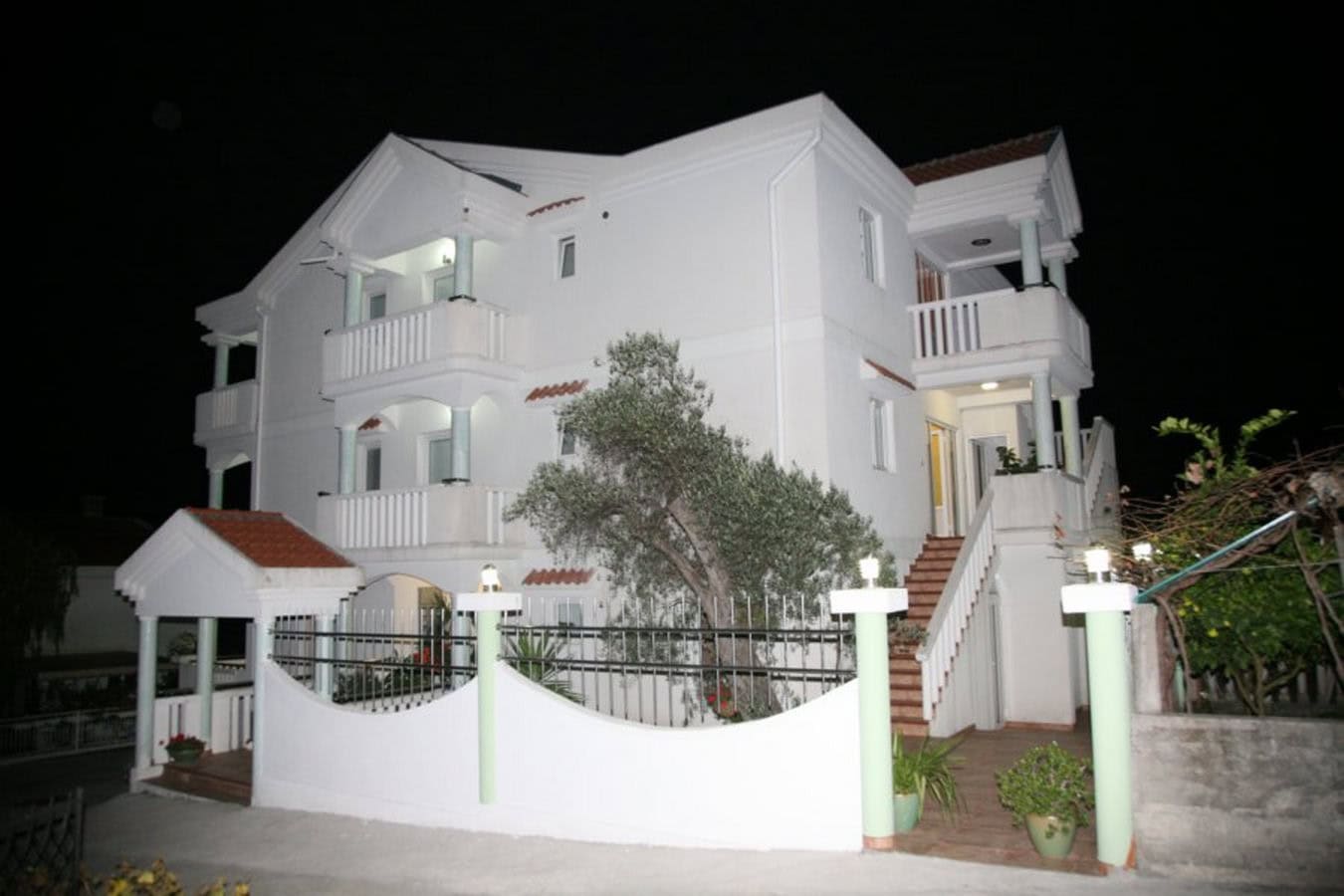Three-story white villa in Tivat, Montenegro, with terraces, an exterior staircase, and fencing at night.