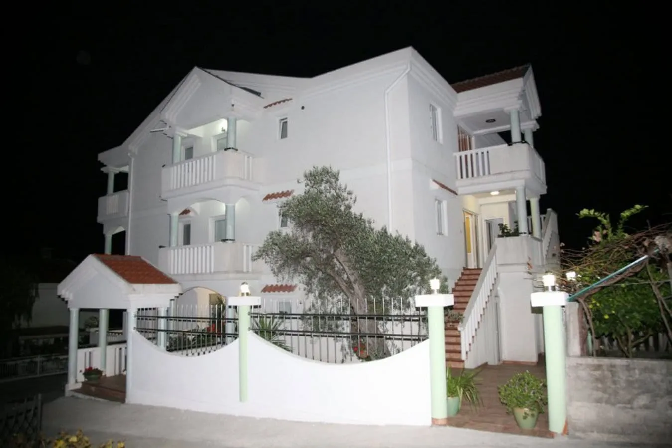 Night view of a white two-story house with balconies, fence, and trees in the foreground.