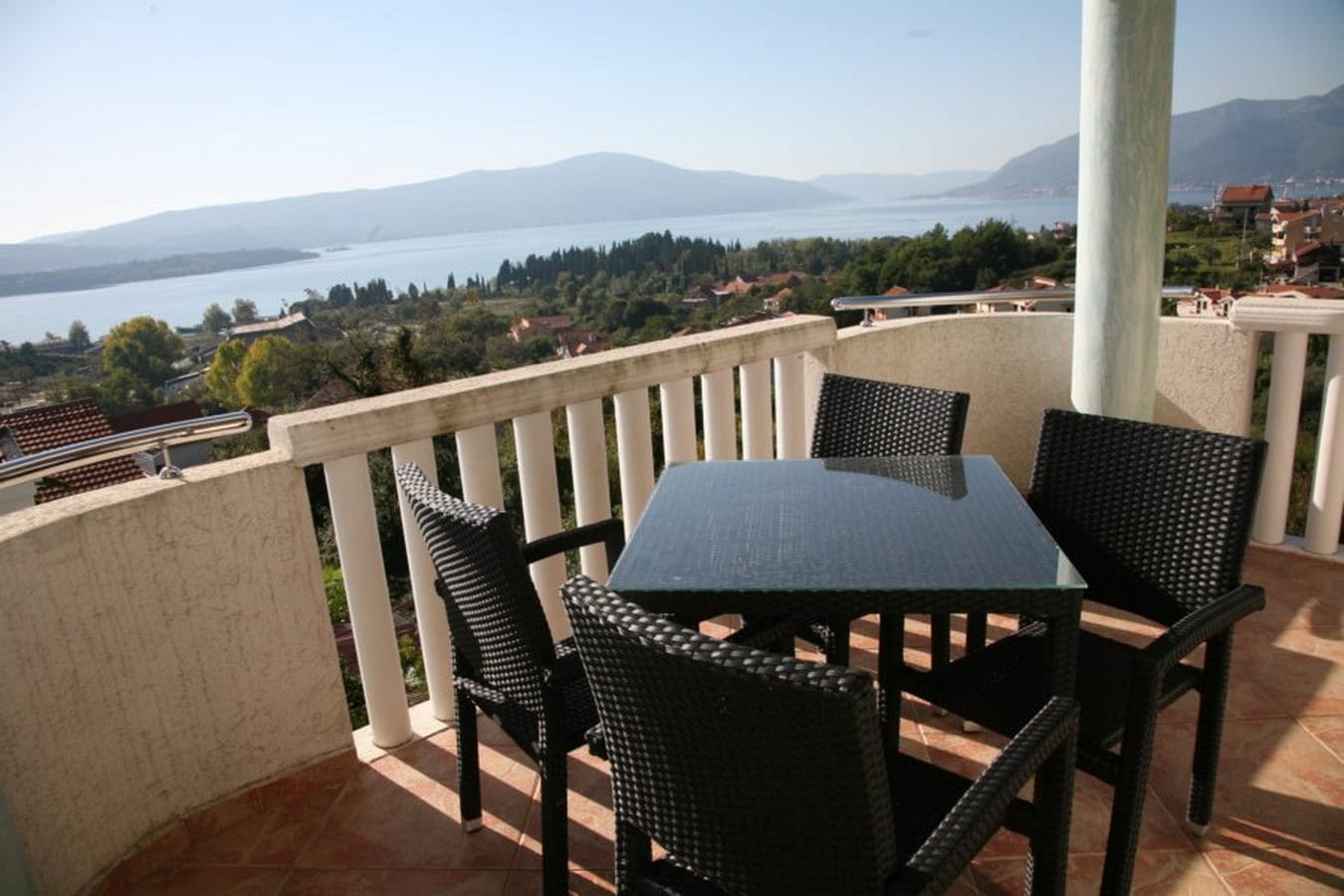 Villa terrace in Tivat, Montenegro: wicker furniture, a glass table, and panoramic views of the Bay of Kotor and the mountains.
