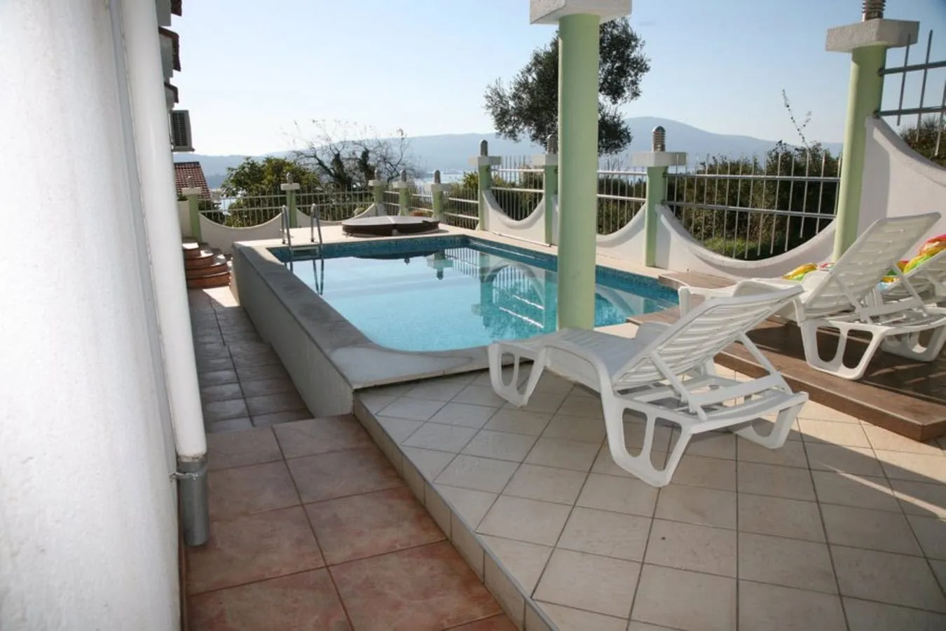 Outdoor pool with white loungers on a tiled terrace overlooking mountains and water.