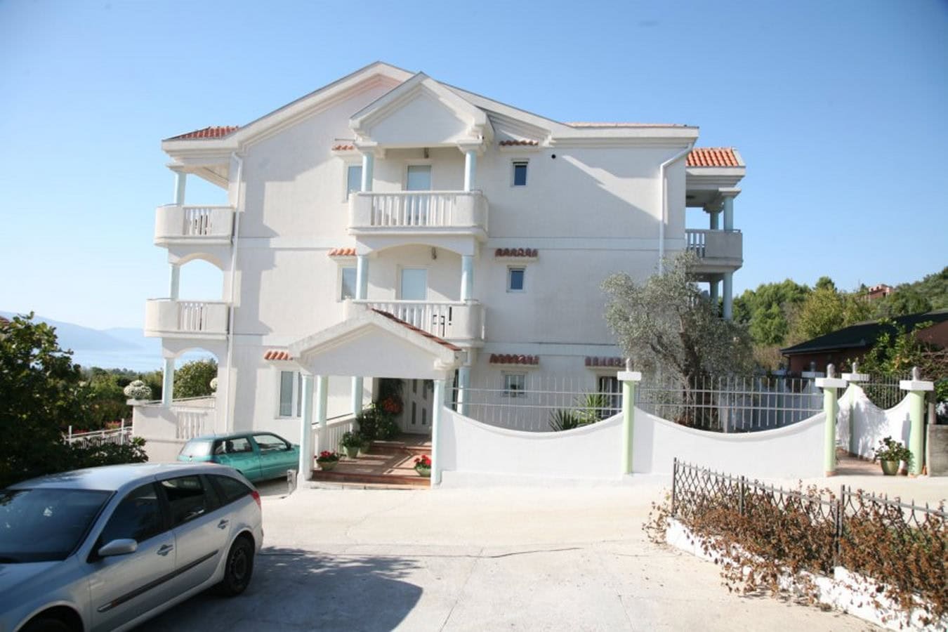 Three-story white villa with balconies and parking in Tivat, Montenegro. Front view of the house with the surrounding grounds.