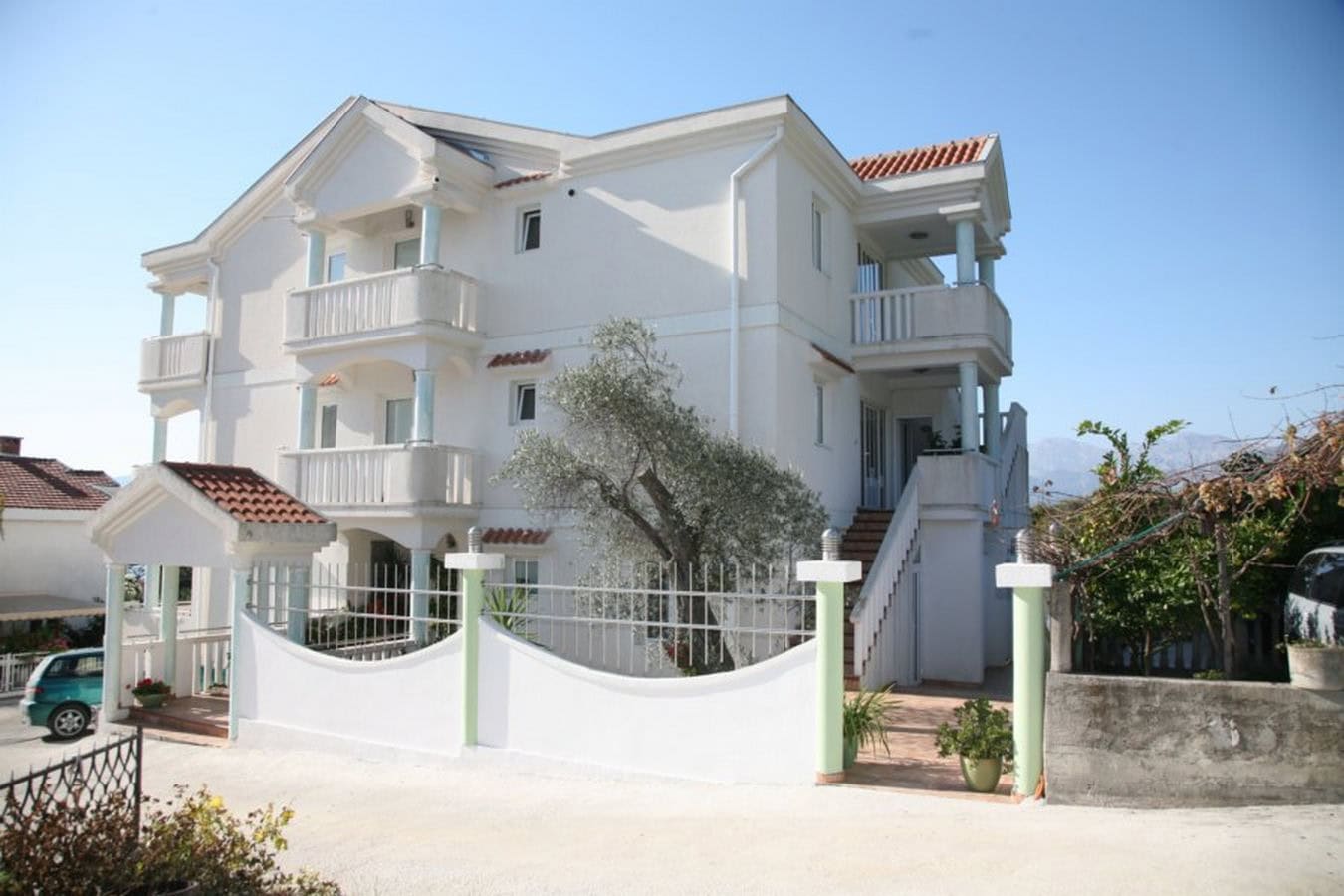 Three-story white villa with balconies and a terrace in Tivat, Montenegro. Facade with a tiled roof and an olive tree.