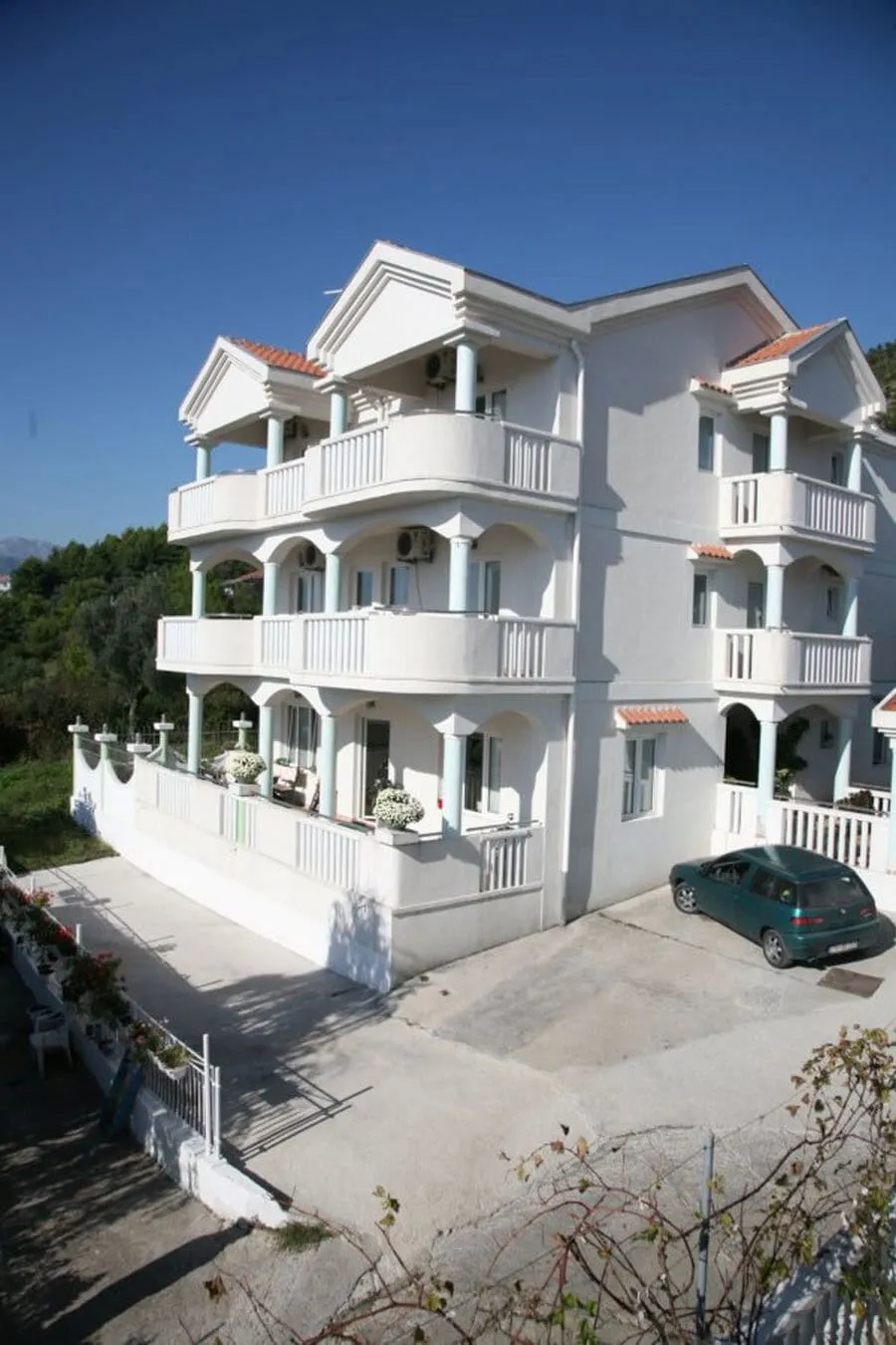 Exterior view of a white multi-story building with balconies, columns, and a green car parked outside under blue sky.
