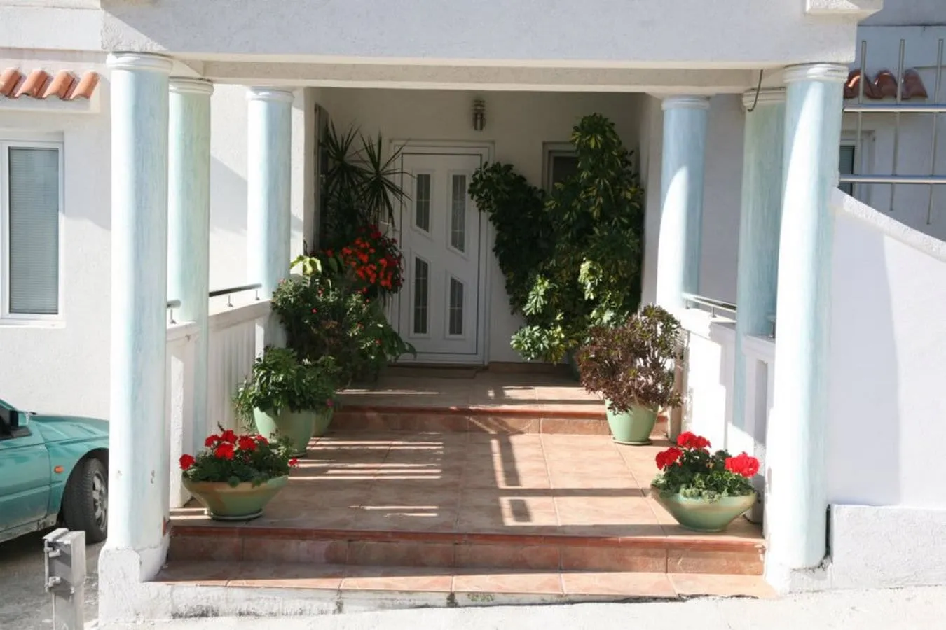 House entrance featuring white columns, terracotta tiled steps, and numerous potted plants with red flowers leading to the front door.
