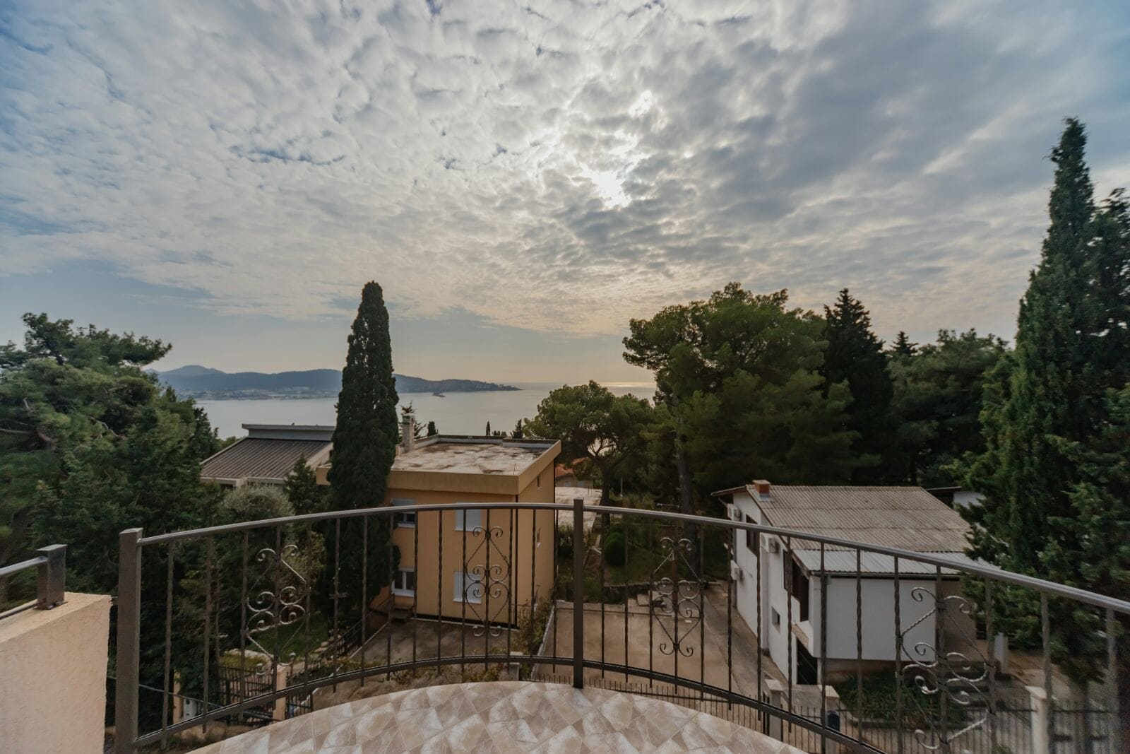 View from the balcony of a house in Shushan, Montenegro: a panorama of the sea, mountains, and cypress trees with a wrought-iron terrace railing.