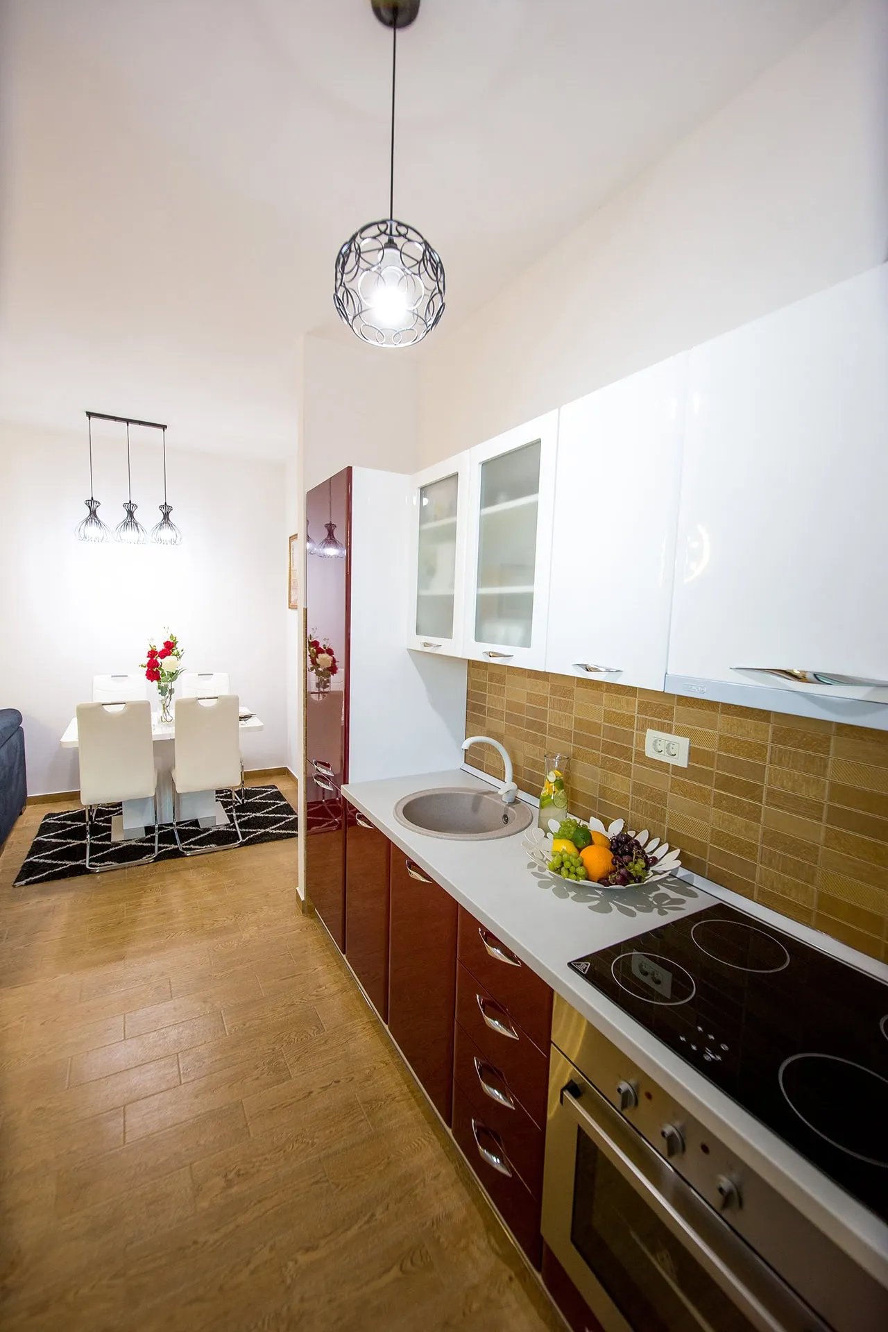 Modern kitchen with red and white cabinets, cooktop, sink, and a dining area visible in the background.