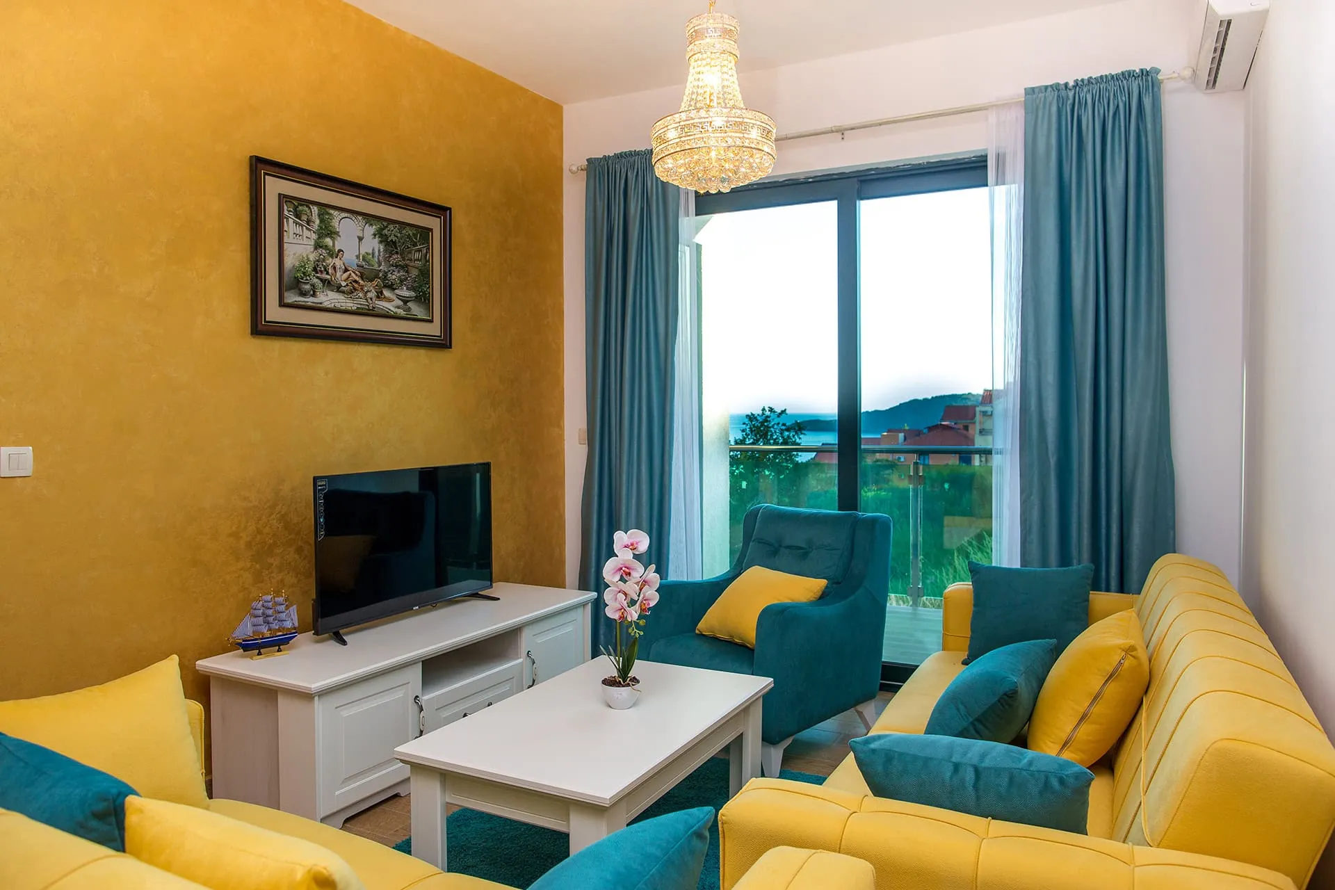 Living room with yellow sofas, blue armchair, TV on white cabinet, and crystal chandelier near window.