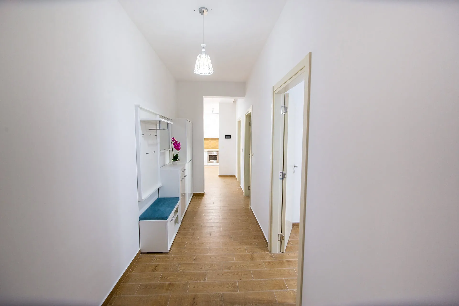 Long white hallway with wood-look tile flooring, a white entryway bench with blue cushion, and open doors.